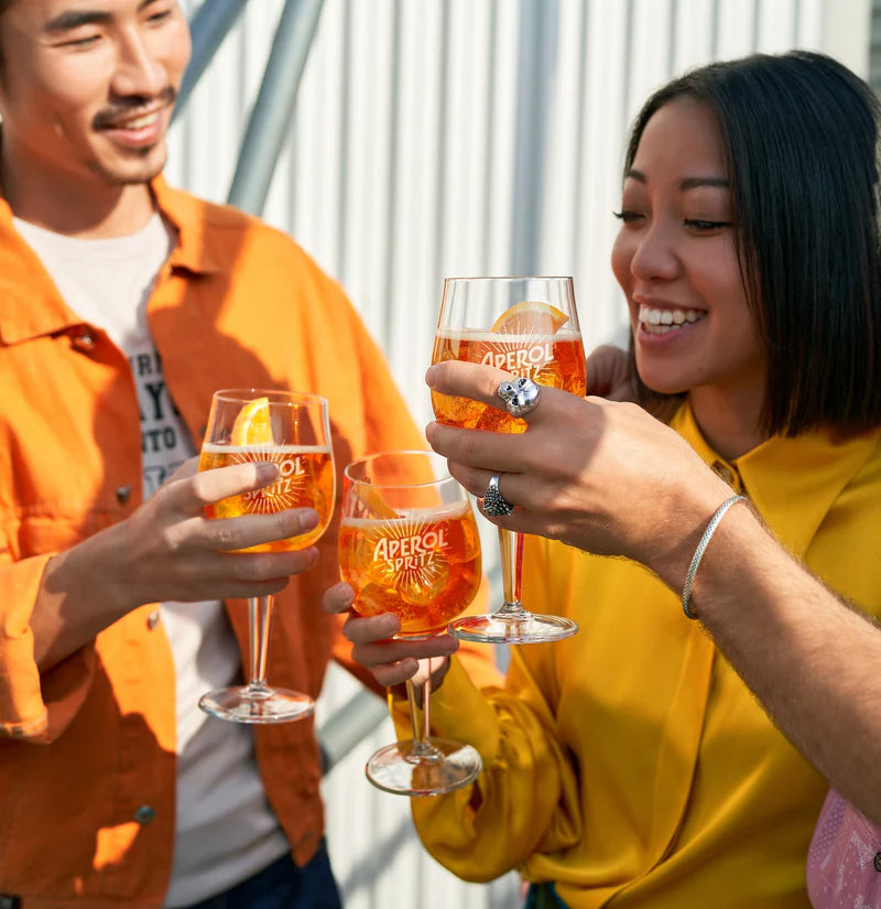 Group of friends clinking Aperol Spritz glasses and smiling outdoors, dressed in vibrant orange and yellow outfits.